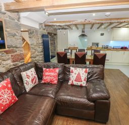 A living room with a brown leather sofa with Christmas cushions next to a kitchen and dining area with chairs at Blacksmiths Cottage in Pembroke