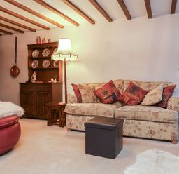 A living room with a patterned sofa and red pillows a wooden cabinet with plates and dolls a floor lamp and a round red chair at Old Farm Cottage in Woolacombe