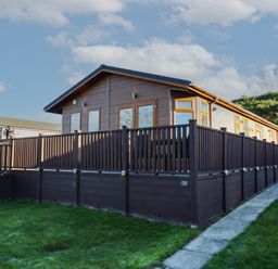 A brown wooden cabin with a fenced deck and grass surrounding it at The Sanctuary Badgers Retreat Holiday Park near Catterick Garrison North Yorkshire