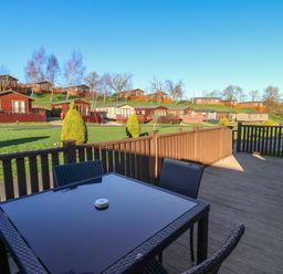 An outdoor patio with a glass table and chairs overlooking wooden lodges on grassy slopes at The Sanctuary Badgers Retreat Holiday Park near Catterick Garrison North Yorkshire