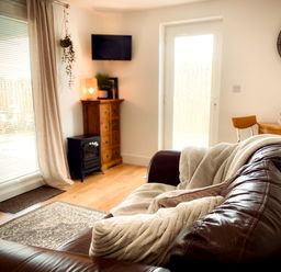 A living room with a leather sofa with cushions and blankets a wooden cabinet a wall-mounted TV and a glass door at The Studio in Crackington Haven
