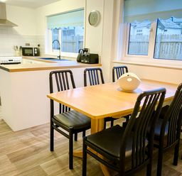 A dining area with a wooden table and six black chairs next to a kitchen with white cabinets and appliances at Trelawne Near Crackington Haven