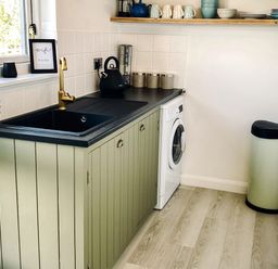 A kitchen area with a black sink gold faucet green cabinets washing machine and shelving with dishes at Penkenna in Crackington Haven