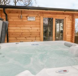 An outdoor hot tub with bubbling water next to a wooden cabin with glass doors at Little Orchard Lodge in Goonhavern