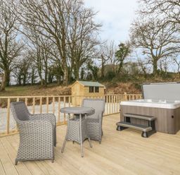An outdoor deck with a hot tub a small table and two chairs and a wooden shed surrounded by leafless trees