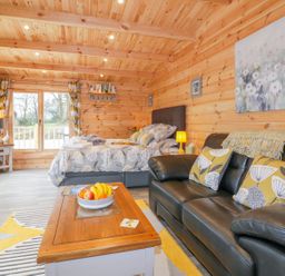 A wooden cabin interior with a bed a black leather sofa a wooden coffee table and large windows at Little Orchard Lodge in Goonhavern