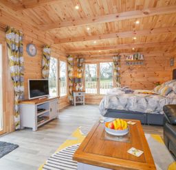 A wooden room with a bed a coffee table with fruit a television and large windows at Little Orchard Lodge in Goonhavern