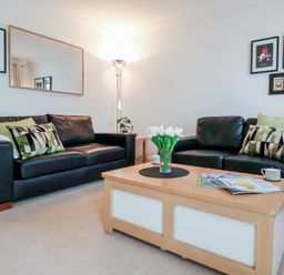 A living room with two black leather sofas a wooden coffee table with magazines and flowers and framed pictures on the wall at Bay Tree Cottage in Peasenhall