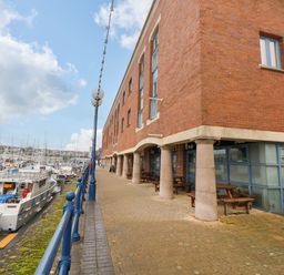 A walkway along a marina with boats docked next to a brick building with columns and outdoor seating at 15 Sovereign House in Milford Haven