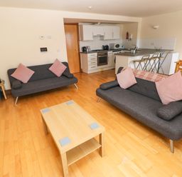 A living room with two sofas pink cushions a wooden coffee table and an open kitchen with bar stools and a dining table at 15 Sovereign House in Milford Haven