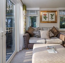 A small sitting area with a sofa, cushions, ottoman, framed botanical prints, windows with blinds, and glass doors leading outside at Lily Rose Lodge in White Cross Bay near Troutbeck Bridge