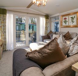 A living room with brown sofas pillows floral curtains and glass doors opening to an outdoor deck at Lily Rose Lodge White Cross Bay near Troutbeck Bridge
