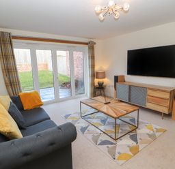 A living room with a gray sofa yellow and gray cushions a wooden coffee table a patterned rug a wooden TV cabinet a wall-mounted TV and sliding glass doors at 4 Centurion Rise in Penrith