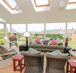 A conservatory with a green sofa pillows wicker chairs wooden rocking chair and large windows at Tree Tops in Newport Pembrokeshire