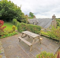 An outdoor patio area with a wooden picnic table surrounded by plants and a stone house in the background at The Farmhouse in Llanbedr