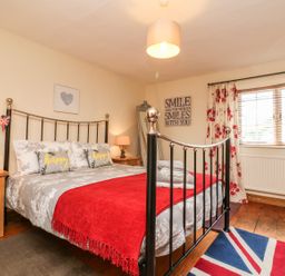 A bedroom with a metal bed frame red blanket bedside tables with lamps and a window with floral curtains at Little Dragons Dunster near Minehead