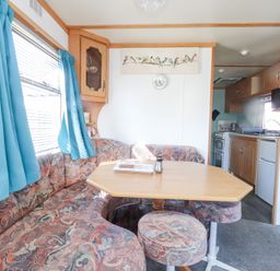 A dining area with patterned seating and a wooden table next to a window with blue curtains and a view into a kitchen at Tanyfron Holiday Caravan in Bryngwran near Gwalchmai