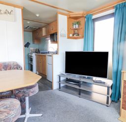 A living room with a wooden dining table and upholstered chairs a television on a stand and a view into a kitchen at Tanyfron Holiday Caravan in Bryngwran near Gwalchmai