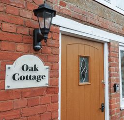 The outside wall of a brick house with a wooden door a black lantern and a sign saying Oak Cottage at Oak Cottage in Aylesbeare