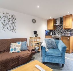 A living room with a brown sofa and blue armchair next to a kitchen with wooden cabinets at Oak Cottage in Aylesbeare