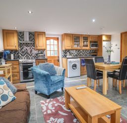An open kitchen and living area with wooden cabinets a blue armchair a brown sofa and a dining table at Oak Cottage in Aylesbeare