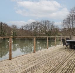 A wooden deck with glass railing overlooking a river with trees and outdoor table and chairs at 2 Millers Island Near Lanreath