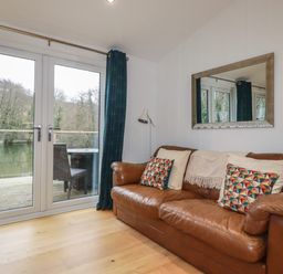 A living room with a brown leather sofa, patterned cushions, a floor lamp, glass doors leading to a balcony overlooking a body of water at 2 Miller's Island Near Lanreath