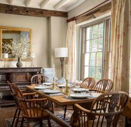 A dining room with wooden table and chairs set near a large window with curtains and an ornate sideboard with a mirror