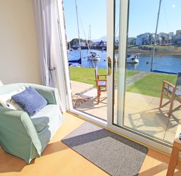 A room with a green armchair and blue cushions next to glass sliding doors opening to a patio with wooden chairs and a water view at Harbour Escape in Porthmadog