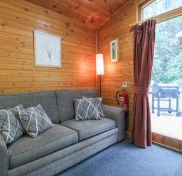 A wooden room with a grey sofa and three cushions next to a floor lamp and glass door with red curtains at Oak Lodge in Rosliston