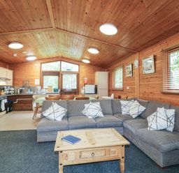 A wooden lodge interior with a gray sectional sofa white patterned cushions wooden coffee table kitchen area and dining table with chairs at Chestnut Lodge in Rosliston