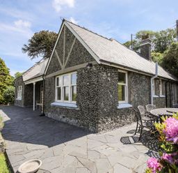 An exterior view of a stone house with a slate roof and a patio with outdoor table and chairs surrounded by plants and trees at Bryn Awelon in Harlech