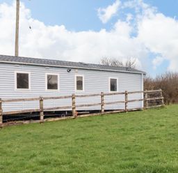 An exterior view of a light blue caravan with four windows and a wooden railing on a grassy hill at Little Trevorrick Caravan in St Issey