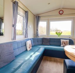 A seating area with blue cushioned benches and a round wooden table by windows with striped curtains and flowers at Little Trevorrick Caravan in St Issey