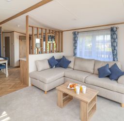 A living room with a beige sectional sofa and blue cushions next to a wooden coffee table with drinks and pastries and a dining table with blue chairs and a flower vase at Maple Lodge in Whitby