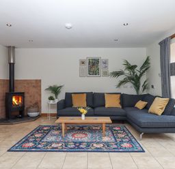 A living room with a dark sectional sofa and a coffee table on a patterned rug near a wood burning stove at The Old Coach House Little Dunley Cottages near Bovey Tracey