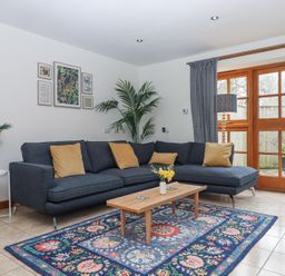 A living room with a blue sectional sofa with yellow cushions a wooden coffee table on a colorful patterned rug plants and framed pictures on the wall at The Old Coach House Little Dunley Cottages near Bovey Tracey