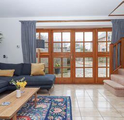A living room with a blue sectional sofa yellow pillows a wooden coffee table with flowers a patterned rug near wooden framed glass doors and carpeted stairs at The Old Coach House Little Dunley Cottages near Bovey Tracey
