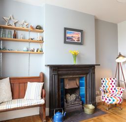 A sitting area with a wooden bench, a fireplace with blue tiles, a colorful armchair, and a floor lamp at School House in Aberffraw