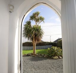 A view through an arched doorway showing a gravel area, a tree, a lawn, and a fence at School House in Aberffraw