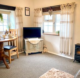 A living room with a round wooden table set for tea two wooden chairs a television on a cabinet and a small stove at Hawthorn in Crackington near Crackington Haven