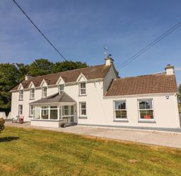 A two-story white farmhouse with a brown tiled roof and porch surrounded by grass at Dromclough Farmhouse in Dromclough near Listowel County Kerry