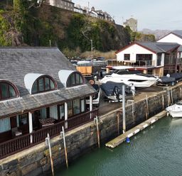 A waterfront scene with boats docked near buildings and a rocky cliff in the background at Fisherman Cove in Porthmadog