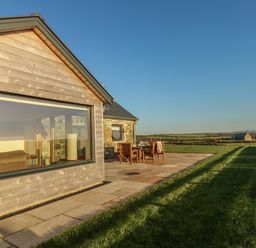 An outdoor stone patio with a wooden table and chairs next to a modern building and grass at Cuddfan in Llanfwrog near Bodedern