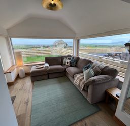 A living room with a sectional sofa and a lamp next to large windows showing a fenced yard at Cuddfan in Llanfwrog near Bodedern