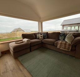 A corner seating area with a brown sofa and cushions near large windows showing a rural view at Cuddfan in Llanfwrog near Bodedern