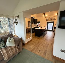 A living area with a sofa and cushions next to a window and an open kitchen with an island and bar stools at Cuddfan in Llanfwrog near Bodedern