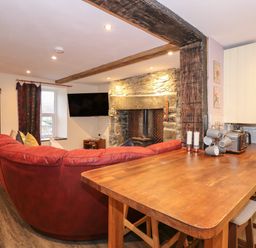 A living room with a red sectional sofa a mounted television a stone fireplace and a wooden kitchen counter with stools at Miners Rest in Blaenau Ffestiniog