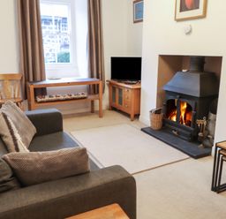 A living room with a grey sofa a wood burning stove a wooden chair and tables at Ty Glyn in Beddgelert