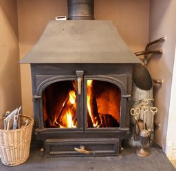 A lit wood-burning stove with a basket of newspapers on the left and fireplace tools on the right at Ty Glyn in Beddgelert